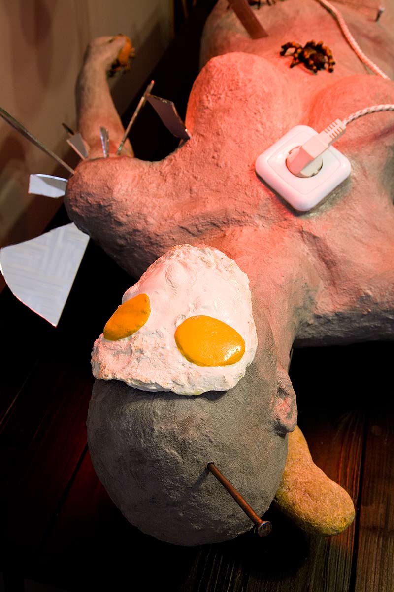 A human figure lies on a table and is being tortured with knives, nails, a fried egg, an iron, scorpions, spiders, broken glass and a vice. Two red lamps illuminate the figure.