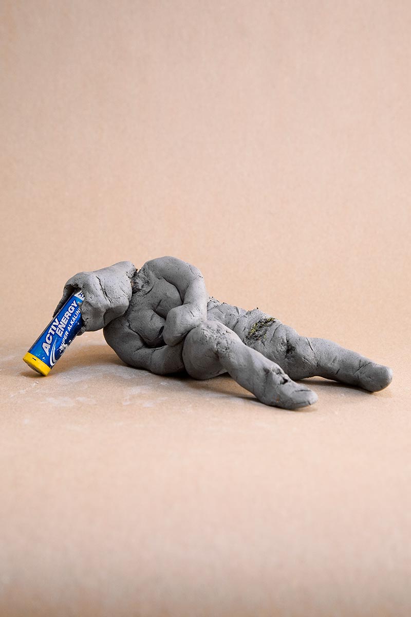 A clay figure lies slumped on the ground. A battery sticks out of its open head.