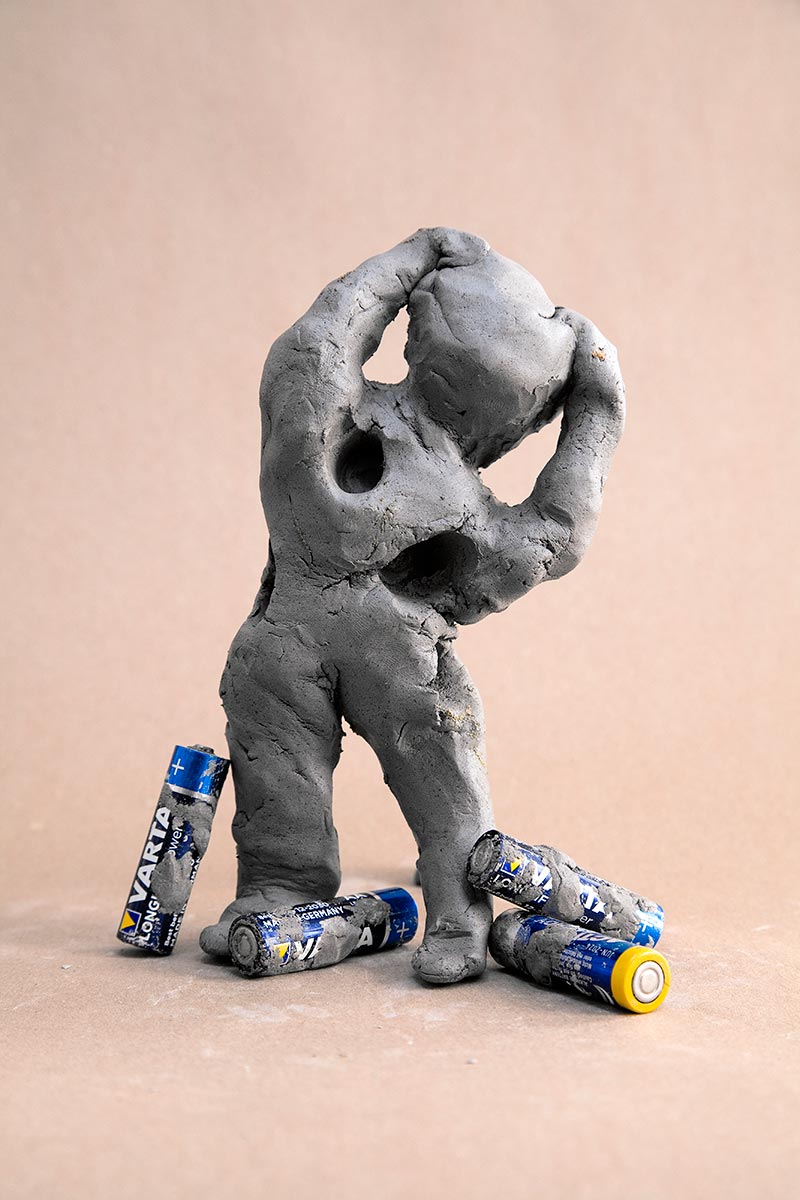 A standing clay figure is desperately holding its head with both arms. Four batteries lie on the ground. In the body of the figure there are holes in which the batteries are previously inserted.